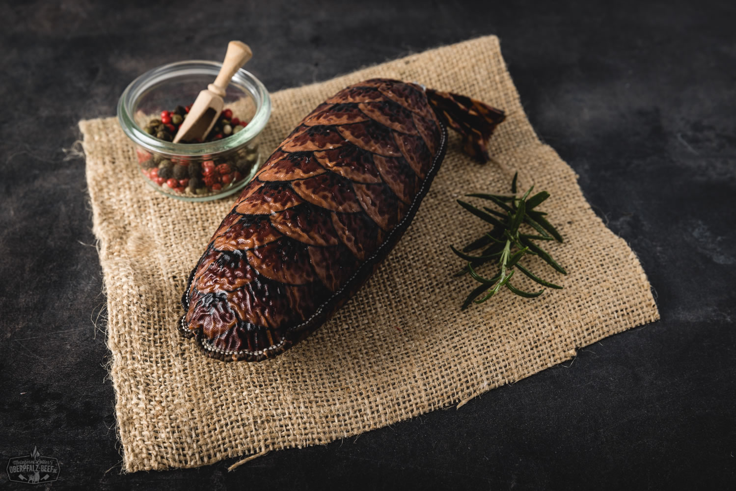 Bild zeigt eine Tannenzapfensalami im Vordergrund, auf traditionellem Holzbrett präsentiert, umgeben von natürlichen Zutaten und Tannenzapfen – rustikale Delikatesse aus der Oberpfalz, perfekt für Feinschmecker und Liebhaber regionaler Fleischprodukte.