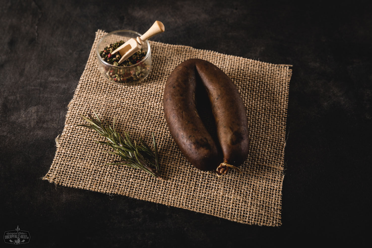 Ein geräucherter Ring Oberpfälzer Leberwurst auf einem rustikalen Holzbrett mit einem Messer, umgeben von frischen Kräutern und Gewürzen.